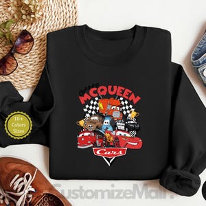 A black sweatshirt with a graphic of the Disney Pixar Cars characters, including Lightning McQueen, is displayed on a white surface.