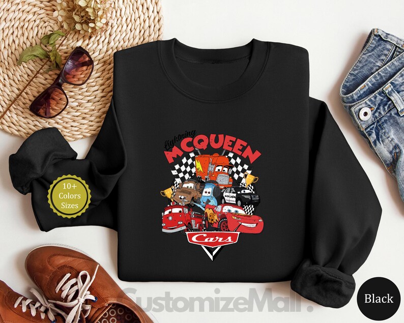 A black sweatshirt with a graphic of the Disney Pixar Cars characters, including Lightning McQueen, is displayed on a white surface.