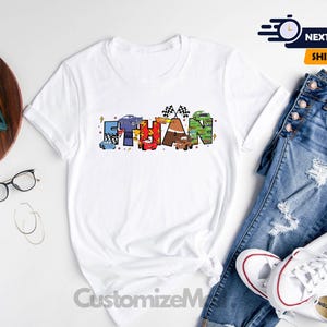 以下が含まれることがあります： 「Ethan」という名前が、漫画の車とレースの旗で構成されたカラフルなグラフィックが描かれた白いTシャツです。