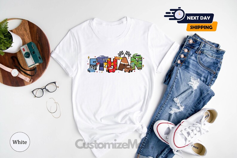 以下が含まれることがあります： 「Ethan」という名前が、漫画の車とレースの旗で構成されたカラフルなグラフィックが描かれた白いTシャツです。