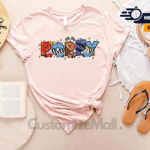 Puede incluir: Una camiseta rosa con un gr&aacute;fico de la palabra "Poppy" formada por coches de dibujos animados. Los coches son rojos, azules y amarillos. La camiseta est&aacute; hecha de un material suave y c&oacute;modo.