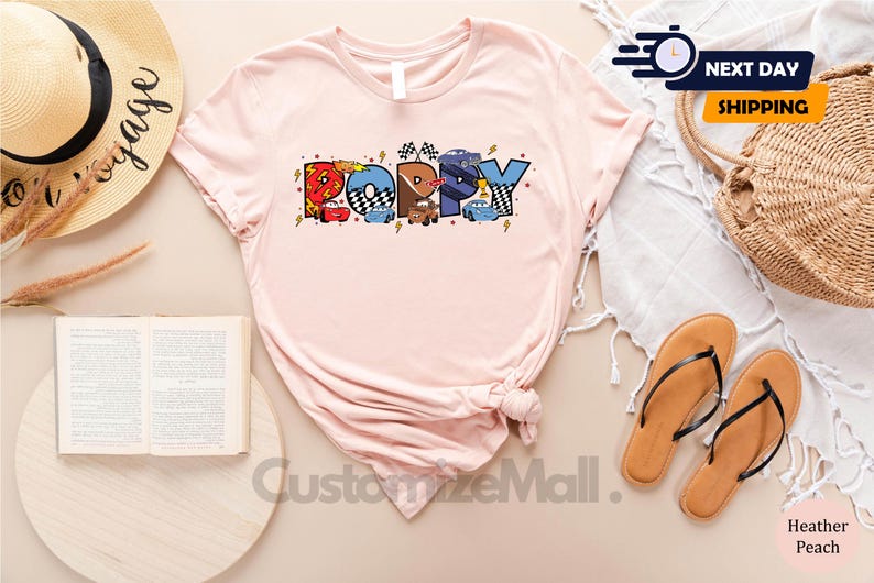 Puede incluir: Una camiseta rosa con un gr&aacute;fico de la palabra "Poppy" formada por coches de dibujos animados. Los coches son rojos, azules y amarillos. La camiseta est&aacute; hecha de un material suave y c&oacute;modo.