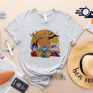 Puede incluir: Camiseta gris claro con una ilustraci&oacute;n de dibujos animados de tres personajes disfrazados para Halloween, con un &aacute;rbol marr&oacute;n y murci&eacute;lagos en el fondo. La camiseta se muestra con accesorios de viaje.