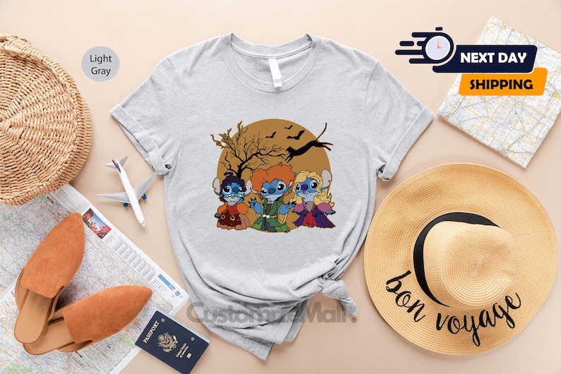 Puede incluir: Camiseta gris claro con una ilustraci&oacute;n de dibujos animados de tres personajes disfrazados para Halloween, con un &aacute;rbol marr&oacute;n y murci&eacute;lagos en el fondo. La camiseta se muestra con accesorios de viaje.