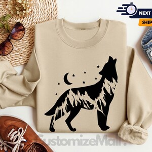 K&ouml;nnte beinhalten: Sandfarbenes Sweatshirt mit einer schwarzen Silhouette eines heulenden Wolfes und einem Bergketten-Design. Das Design beinhaltet einen Mond und Sterne. Das Sweatshirt ist ein Rundhals-Stil.