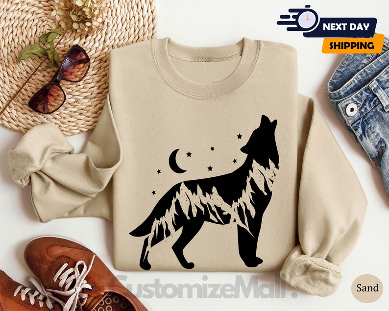 K&ouml;nnte beinhalten: Sandfarbenes Sweatshirt mit einer schwarzen Silhouette eines heulenden Wolfes und einem Bergketten-Design. Das Design beinhaltet einen Mond und Sterne. Das Sweatshirt ist ein Rundhals-Stil.