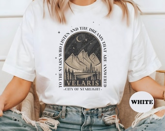 May include: White t-shirt featuring a mountain and city landscape design with the text "VELARIS CITY OF STARLIGHT". The shirt also includes the text "TO THE STARS WHO LISTEN AND THE DREAMS THAT ARE ANSWERED".