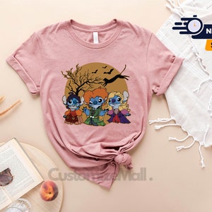 Puede incluir: Camiseta color malva claro con una ilustraci&oacute;n de dibujos animados de tres personajes de Stitch vestidos como las hermanas Sanderson de Hocus Pocus. El dise&ntilde;o est&aacute; sobre un c&iacute;rculo marr&oacute;n con un &aacute;rbol y murci&eacute;lagos. La camiseta tiene mangas enrolladas.