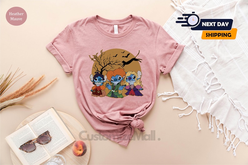 Puede incluir: Camiseta color malva claro con una ilustraci&oacute;n de dibujos animados de tres personajes de Stitch vestidos como las hermanas Sanderson de Hocus Pocus. El dise&ntilde;o est&aacute; sobre un c&iacute;rculo marr&oacute;n con un &aacute;rbol y murci&eacute;lagos. La camiseta tiene mangas enrolladas.