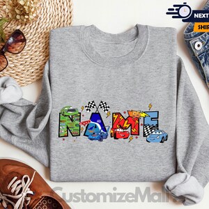 以下が含まれることがあります： グレーのトレーナーに、漫画のレーシングカーで「NAME」という単語がカラフルに描かれています。車は赤、青、緑、黄色の4色です。グラフィックには、市松模様の旗と稲妻も含まれています。