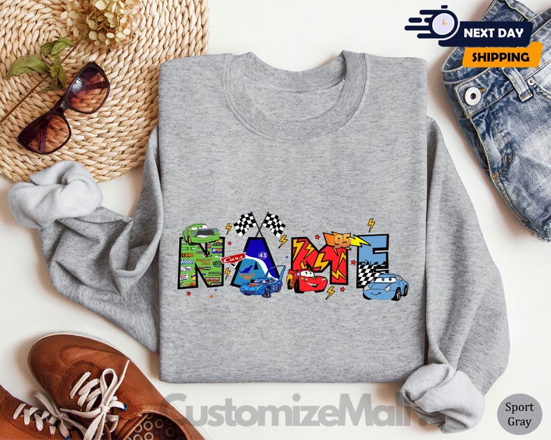 以下が含まれることがあります： グレーのトレーナーに、漫画のレーシングカーで「NAME」という単語がカラフルに描かれています。車は赤、青、緑、黄色の4色です。グラフィックには、市松模様の旗と稲妻も含まれています。