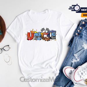 Puede incluir: Camiseta blanca con un dise&ntilde;o gr&aacute;fico colorido que dice "T&iacute;o" con coches de dibujos animados y banderas de carreras.