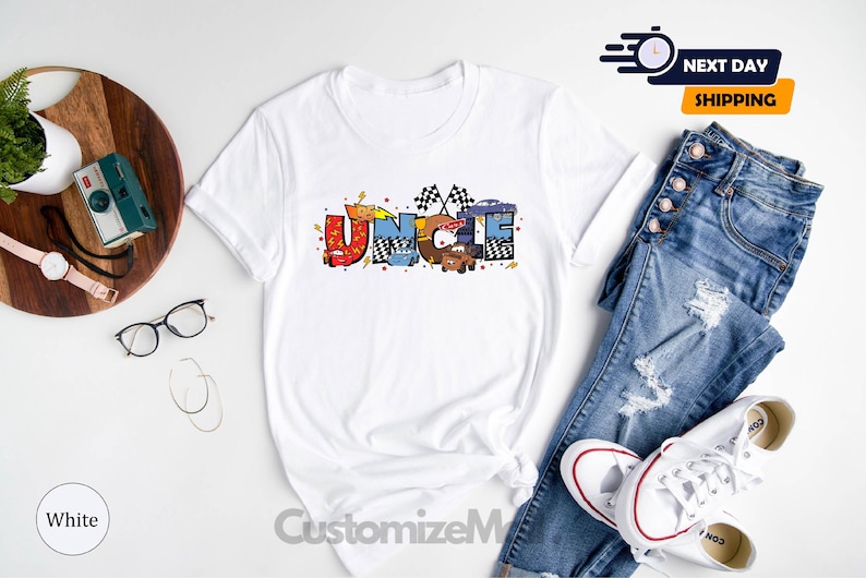 Puede incluir: Camiseta blanca con un dise&ntilde;o gr&aacute;fico colorido que dice "T&iacute;o" con coches de dibujos animados y banderas de carreras.