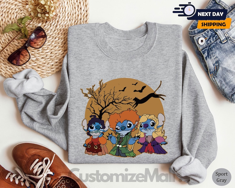 Puede incluir: Una sudadera gris con un dise&ntilde;o tem&aacute;tico de Halloween. El dise&ntilde;o presenta tres personajes de dibujos animados disfrazados de brujas frente a un c&iacute;rculo marr&oacute;n con un &aacute;rbol, murci&eacute;lagos y un gato negro. El texto "CustomizeMall" est&aacute; en la parte inferior.