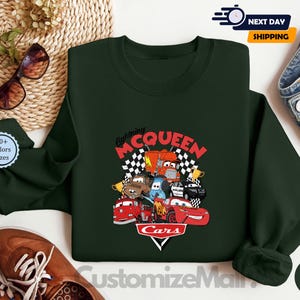 a green sweatshirt with a colorful design of characters from the Cars movie franchise, including Lightning McQueen, Lightning McQueen's car, and other iconic vehicles. The sweatshirt is displayed on a white background, along with a pair of sunglasses, a pair of jeans, and a pair of brown shoes.