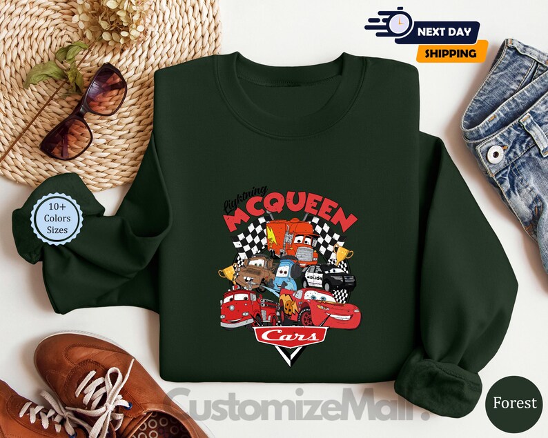 a green sweatshirt with a colorful design of characters from the Cars movie franchise, including Lightning McQueen, Lightning McQueen's car, and other iconic vehicles. The sweatshirt is displayed on a white background, along with a pair of sunglasses, a pair of jeans, and a pair of brown shoes.