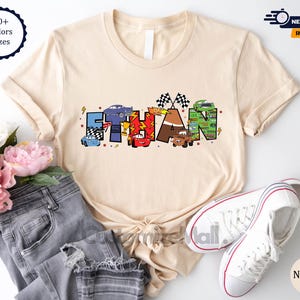 Puede incluir: Una camiseta beige con un dise&ntilde;o tem&aacute;tico de Cars que presenta el nombre "ETHAN" en letras de colores con coches de dibujos animados y banderas de carreras.