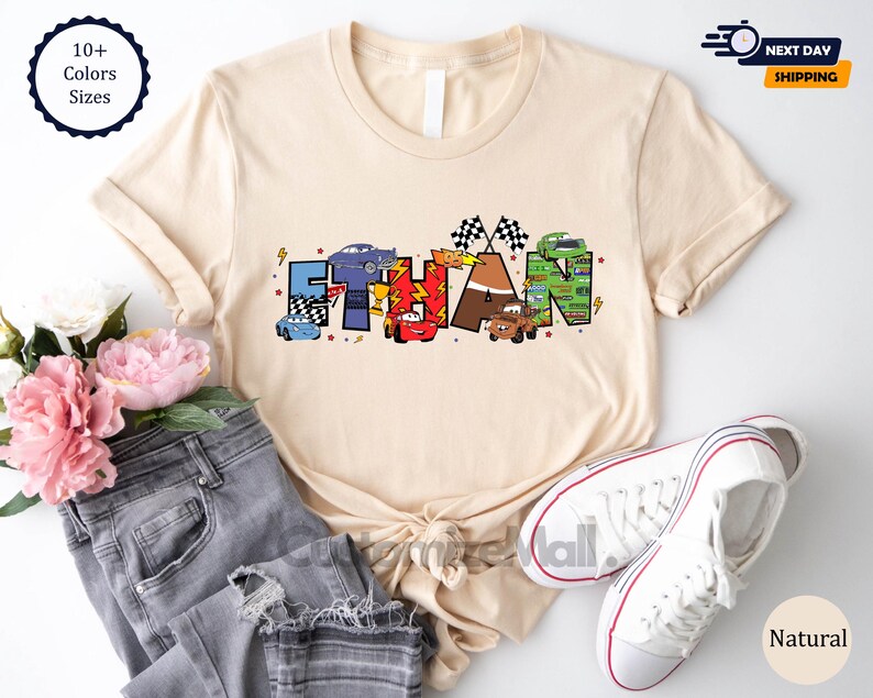 Puede incluir: Una camiseta beige con un dise&ntilde;o tem&aacute;tico de Cars que presenta el nombre "ETHAN" en letras de colores con coches de dibujos animados y banderas de carreras.