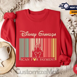 A red sweater with a Disney Grandpa design and the text "Disney Grandpa" printed on it, along with the text "SCAN FOR PAYMENT" below it. The sweater is displayed on a bed with a white blanket and a mug.