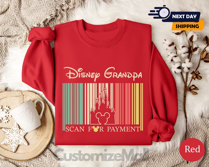 A red sweater with a Disney Grandpa design and the text "Disney Grandpa" printed on it, along with the text "SCAN FOR PAYMENT" below it. The sweater is displayed on a bed with a white blanket and a mug.
