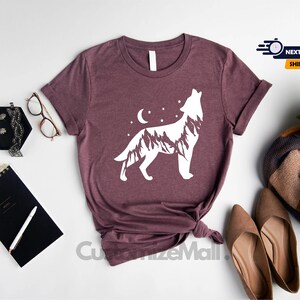 K&ouml;nnte beinhalten: Burgunderfarbenes T-Shirt mit wei&szlig;em Wolfsmotiv, Bergen, Mond und Sternen. Das Shirt wird mit Accessoires wie Brille, Notizbuch und Schuhen pr&auml;sentiert. Der Text "CustomizeMall" ist sichtbar.