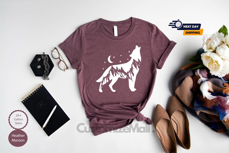 K&ouml;nnte beinhalten: Burgunderfarbenes T-Shirt mit wei&szlig;em Wolfsmotiv, Bergen, Mond und Sternen. Das Shirt wird mit Accessoires wie Brille, Notizbuch und Schuhen pr&auml;sentiert. Der Text "CustomizeMall" ist sichtbar.