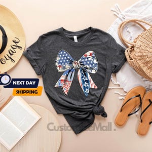 May include: A dark gray t-shirt with a red, white, and blue bow made of American flag fabric. The bow is centered on the shirt.