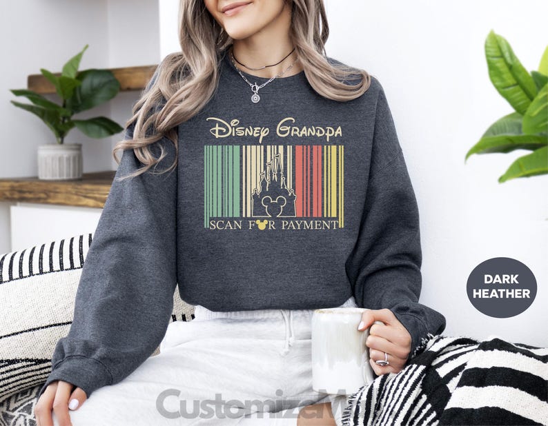 A woman wearing a gray sweatshirt with a Disney Grandpa design is sitting on a couch, holding a mug, and surrounded by potted plants.
