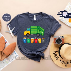 May include: A navy blue t-shirt with a graphic of a green garbage truck and a row of colorful recycling bins. The text "bon voyage" is written on a straw hat in the background.