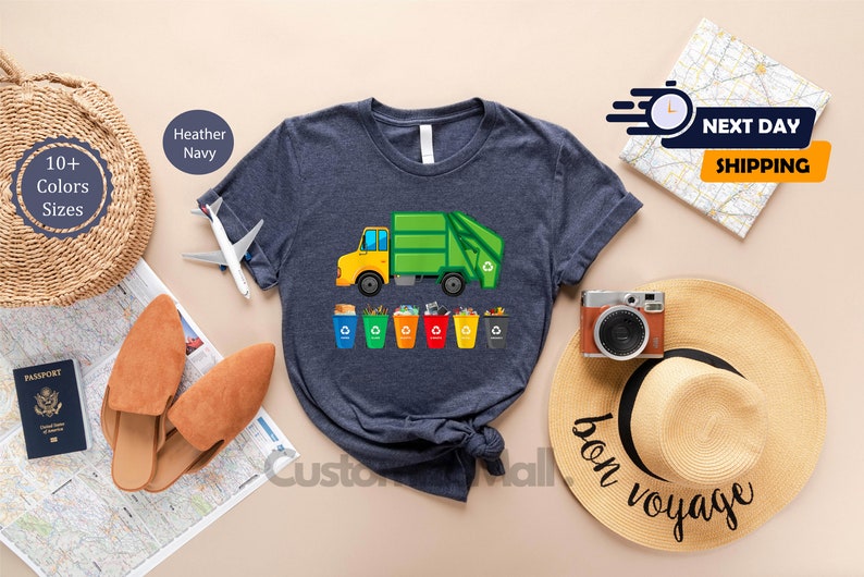 May include: A navy blue t-shirt with a graphic of a green garbage truck and a row of colorful recycling bins. The text "bon voyage" is written on a straw hat in the background.