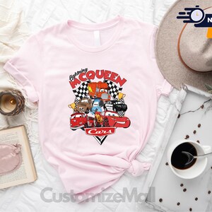 a pink t-shirt with a graphic of characters from the animated film "Cars", along with a hat, a book, a cup of coffee, and other items arranged on a bed.