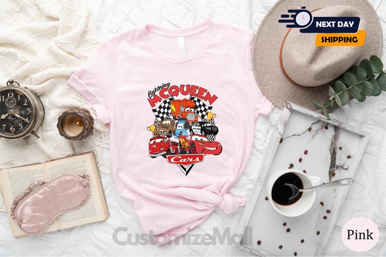 a pink t-shirt with a graphic of characters from the animated film "Cars", along with a hat, a book, a cup of coffee, and other items arranged on a bed.