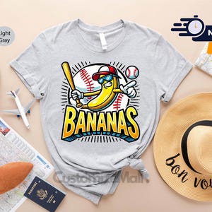 K&ouml;nnte beinhalten: Hellgraues T-Shirt mit einer Cartoon-Banane, die eine Sonnenbrille und eine Baseballkappe tr&auml;gt und einen Schl&auml;ger h&auml;lt. Das Wort "BANANAS" steht in fetten gelben und blauen Buchstaben. Ein Baseball und eine Baseball-Grafik sind ebenfalls im Design enthalten.