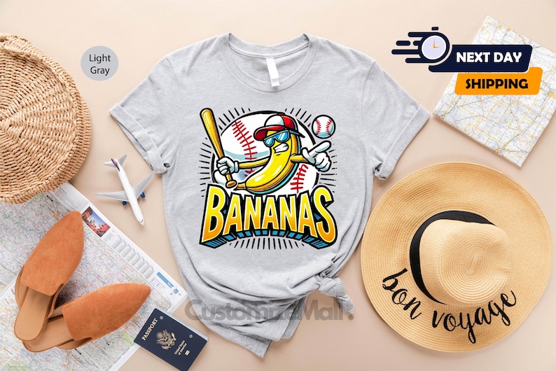 K&ouml;nnte beinhalten: Hellgraues T-Shirt mit einer Cartoon-Banane, die eine Sonnenbrille und eine Baseballkappe tr&auml;gt und einen Schl&auml;ger h&auml;lt. Das Wort "BANANAS" steht in fetten gelben und blauen Buchstaben. Ein Baseball und eine Baseball-Grafik sind ebenfalls im Design enthalten.