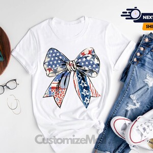 May include: White t-shirt with a red, white, and blue bow design. The bow is made of fabric with a stars and stripes pattern. The shirt is a blank canvas for customization.
