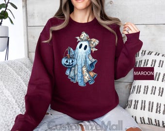 May include: A maroon sweatshirt featuring a watercolor illustration of a ghost holding a pumpkin-shaped basket. The ghost is adorned with a floral bow and patchwork details. The design is set against a plain background.
