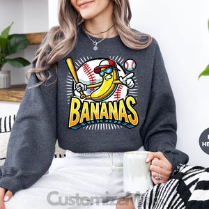 K&ouml;nnte beinhalten: Dunkelgraues Sweatshirt mit einer Cartoon-Banane, die Baseball spielt. Die Banane tr&auml;gt eine Sonnenbrille und eine Baseballkappe und h&auml;lt einen Schl&auml;ger. Das Wort "BANANAS" steht in fetten, farbigen Buchstaben. Ein Baseball ist im Hintergrund.