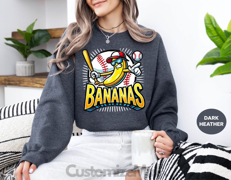 K&ouml;nnte beinhalten: Dunkelgraues Sweatshirt mit einer Cartoon-Banane, die Baseball spielt. Die Banane tr&auml;gt eine Sonnenbrille und eine Baseballkappe und h&auml;lt einen Schl&auml;ger. Das Wort "BANANAS" steht in fetten, farbigen Buchstaben. Ein Baseball ist im Hintergrund.
