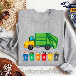 May include: A gray sweatshirt with a green garbage truck and recycling bins printed on it. The truck has a yellow cab and the bins are labeled with different recycling symbols. The text "CustomizeMall" is printed on the bottom of the sweatshirt.