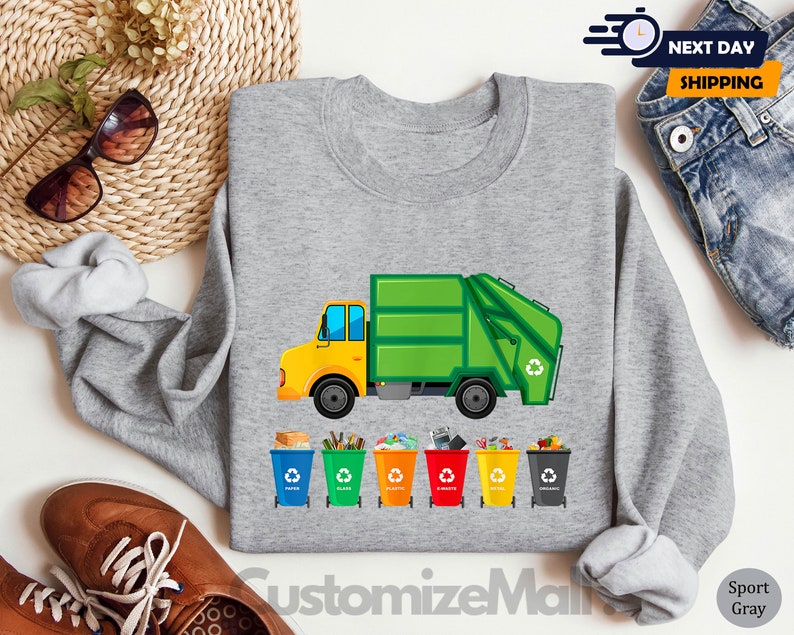 May include: A gray sweatshirt with a green garbage truck and recycling bins printed on it. The truck has a yellow cab and the bins are labeled with different recycling symbols. The text "CustomizeMall" is printed on the bottom of the sweatshirt.