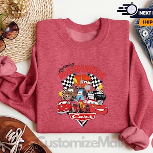 a red sweatshirt with a graphic of various cars and characters from the Cars movie, along with the text "Lightning McQueen". The sweatshirt is placed on a white surface, surrounded by other items such as sunglasses, jeans, and a pair of shoes.