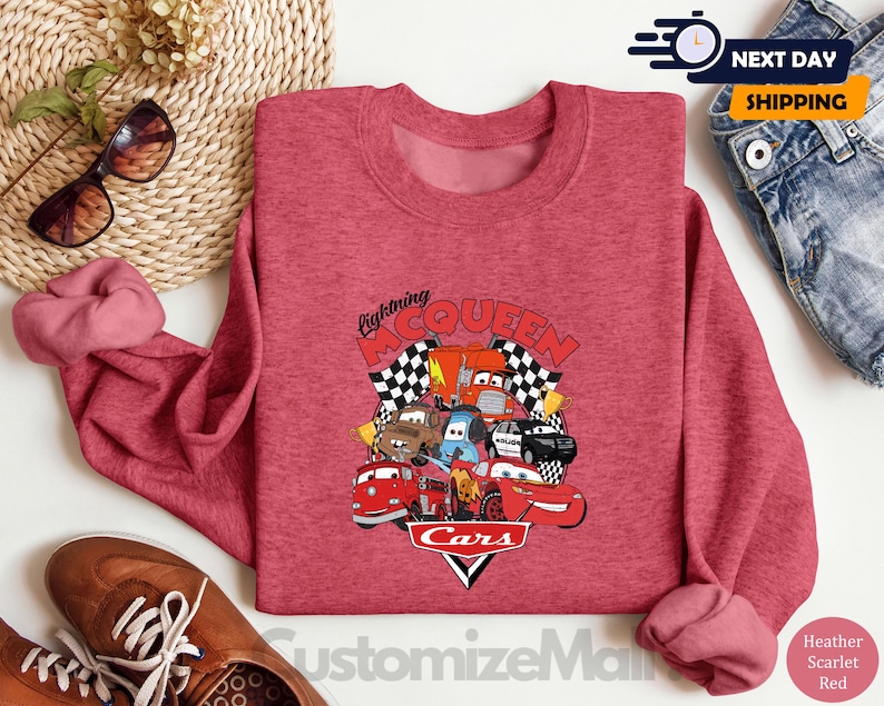 a red sweatshirt with a graphic of various cars and characters from the Cars movie, along with the text "Lightning McQueen". The sweatshirt is placed on a white surface, surrounded by other items such as sunglasses, jeans, and a pair of shoes.