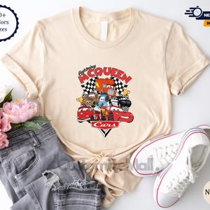 a casual outfit consisting of a beige t-shirt with a graphic of a cartoon car, a pair of jeans, and a pair of white sneakers, along with some pink flowers.