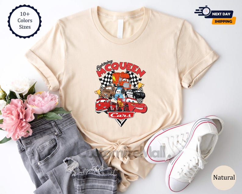 a casual outfit consisting of a beige t-shirt with a graphic of a cartoon car, a pair of jeans, and a pair of white sneakers, along with some pink flowers.