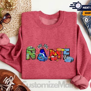 Puede incluir: Sudadera roja de mezcla con un gr&aacute;fico colorido de la palabra "NAME" formado por coches de dibujos animados y banderas de carreras.