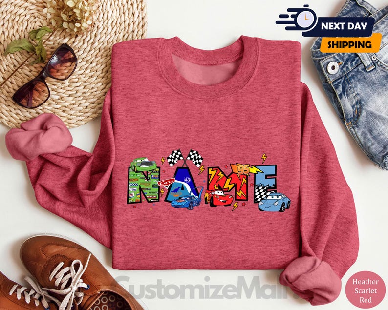 Puede incluir: Sudadera roja de mezcla con un gr&aacute;fico colorido de la palabra "NAME" formado por coches de dibujos animados y banderas de carreras.