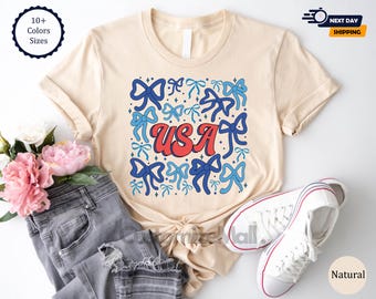 May include: A beige t-shirt with a graphic design of blue and red bows surrounding the word "USA" in red letters. The shirt is available in multiple colors and sizes.