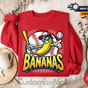 K&ouml;nnte beinhalten: Rotes Sweatshirt mit einer Cartoon-Banane, die eine Sonnenbrille und eine Baseballkappe tr&auml;gt und einen Schl&auml;ger h&auml;lt. Das Design beinhaltet einen Baseball und das Wort "BANANAS" in Gelb und Blau. Das Sweatshirt hat hochgekrempelte &Auml;rmel.
