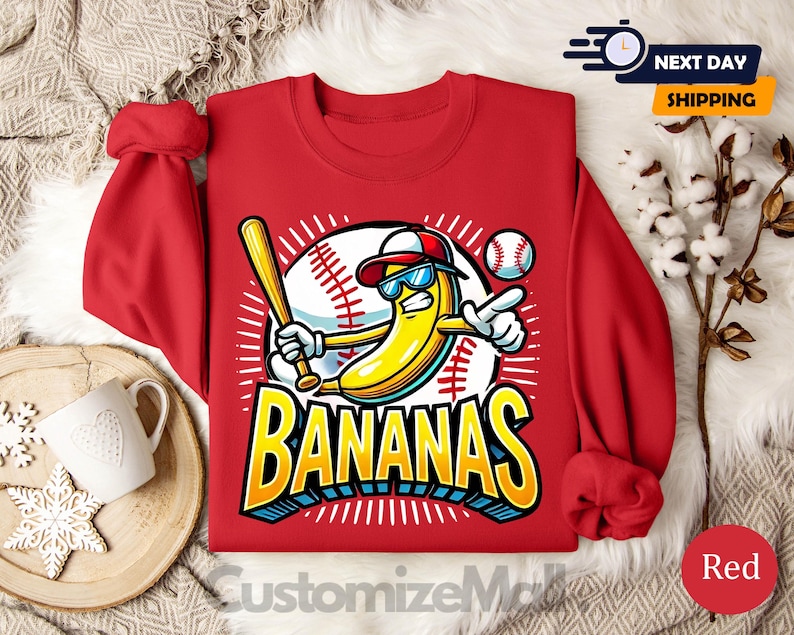 K&ouml;nnte beinhalten: Rotes Sweatshirt mit einer Cartoon-Banane, die eine Sonnenbrille und eine Baseballkappe tr&auml;gt und einen Schl&auml;ger h&auml;lt. Das Design beinhaltet einen Baseball und das Wort "BANANAS" in Gelb und Blau. Das Sweatshirt hat hochgekrempelte &Auml;rmel.