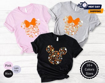 May include: Three t-shirts in pink, light gray, and black, each featuring a Halloween-themed design. The design is a mouse head shape filled with Halloween icons and an orange bow. The shirts are displayed with the words "Pink", "Light Gray", and "Black".